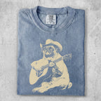 Blue t-shirt with a tan illustration of a dog wearing a cowboy hat playing a guitar.