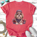 Shih Tzu wearing sunglasses and a USA bandana, seated among patriotic floral decorations.