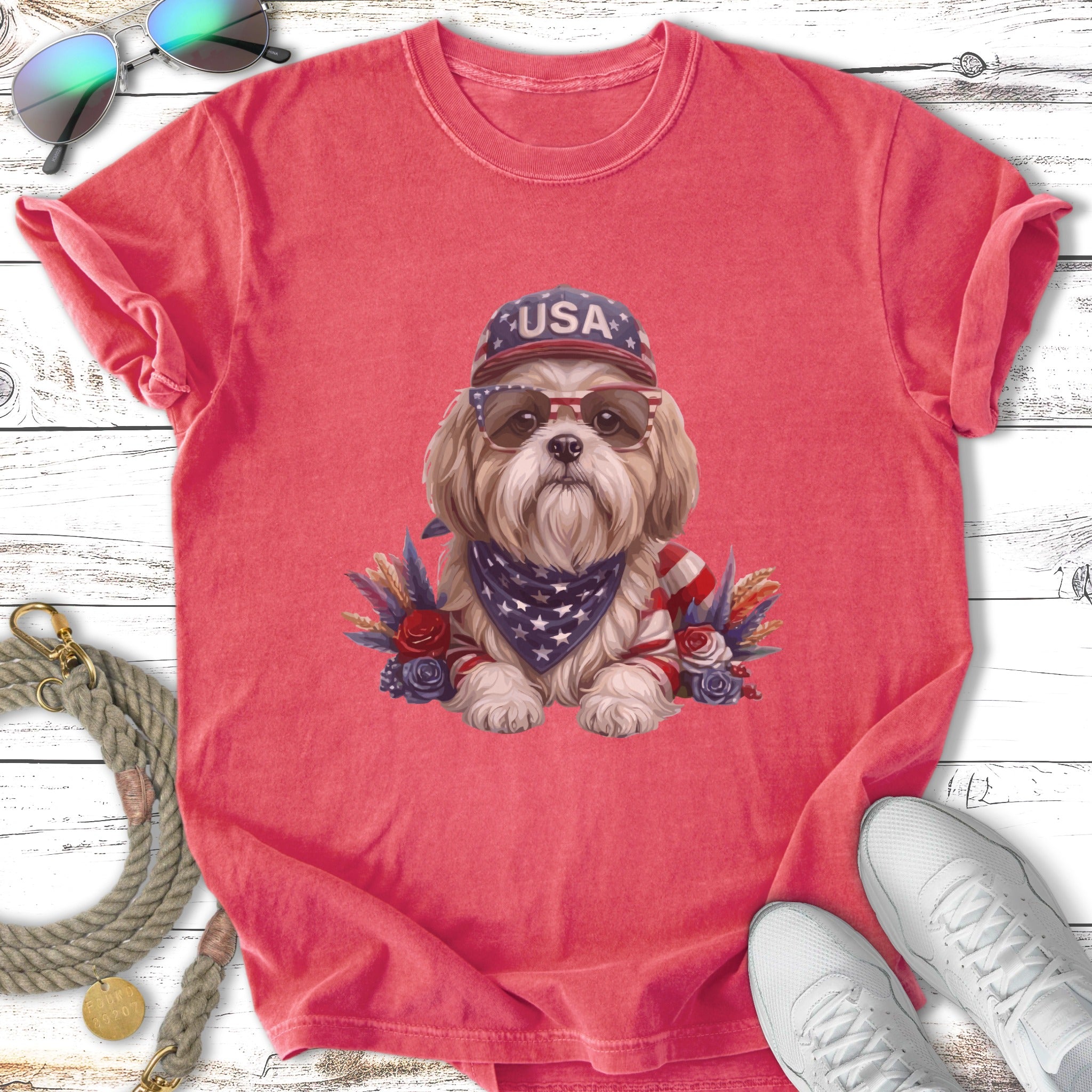 Shih Tzu wearing sunglasses and a USA bandana, seated among patriotic floral decorations.