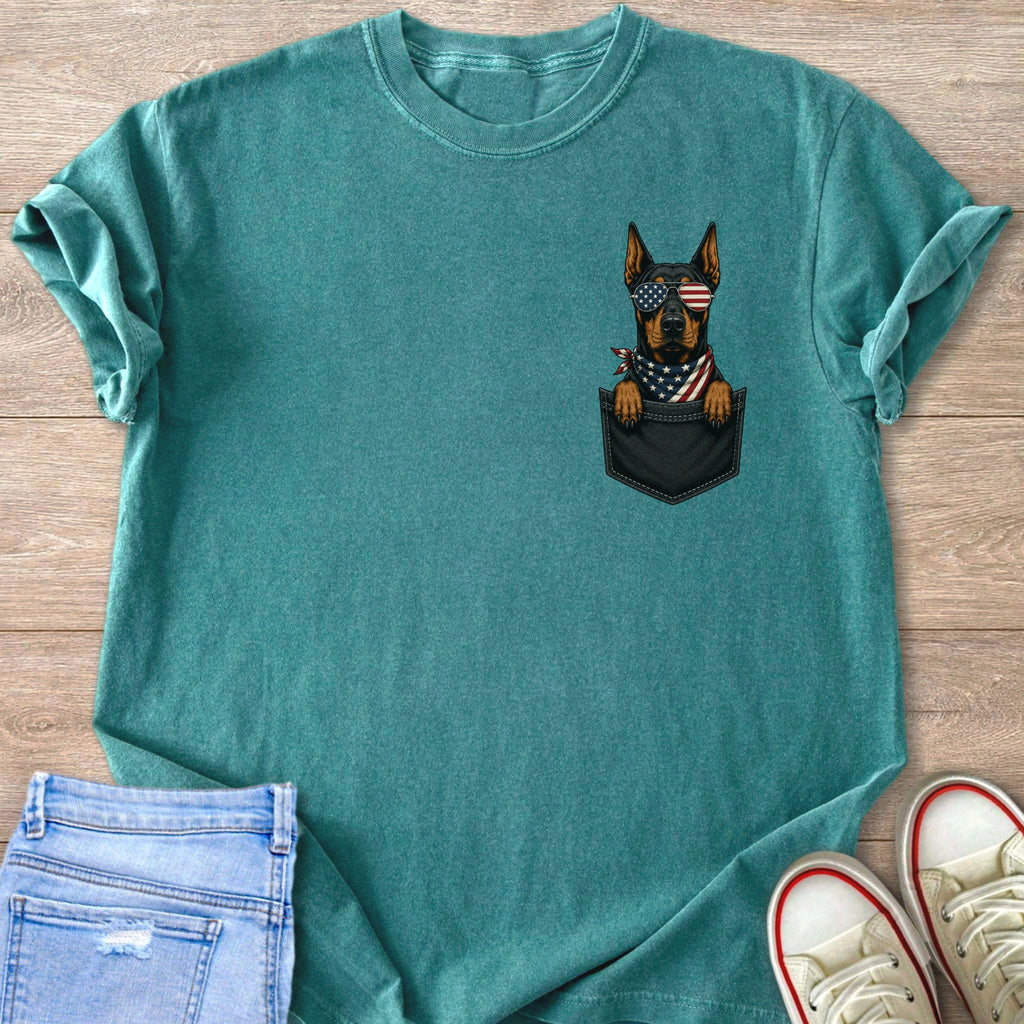 Doberman wearing American flag sunglasses peeking from a pocket on a teal t-shirt.