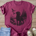 Black Labrador and ducks near water with pine trees, printed on a maroon t-shirt.
