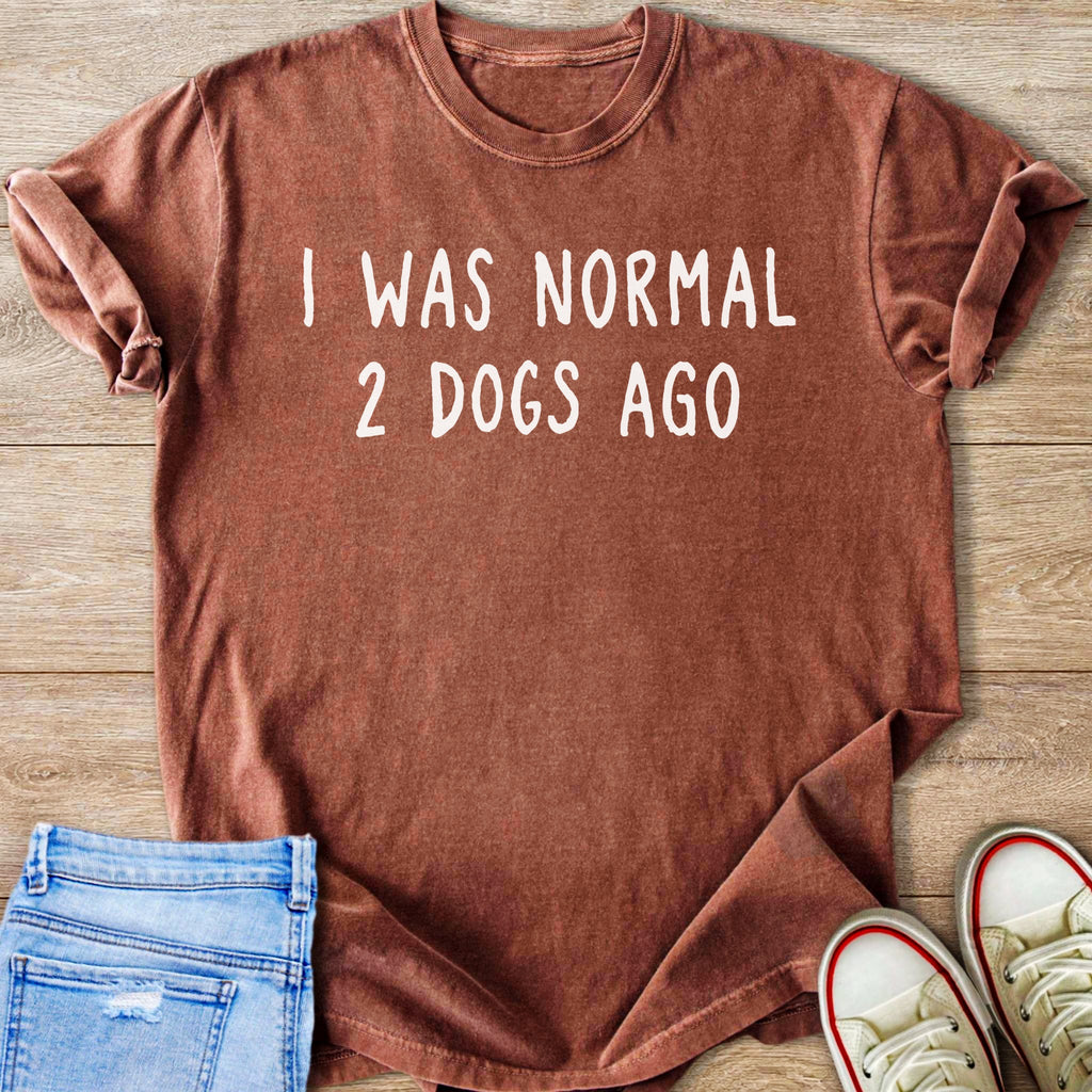 Brown t-shirt with white text "I WAS NORMAL 2 DOGS AGO" on wooden background.