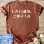Brown t-shirt with white text "I WAS NORMAL 2 DOGS AGO" on wooden background.