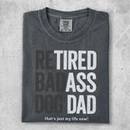 Dark grey t-shirt with "RETIRED BADASS DOG DAD" in large block letters.