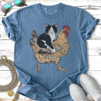 Cat sitting on a chicken graphic on a blue t-shirt.