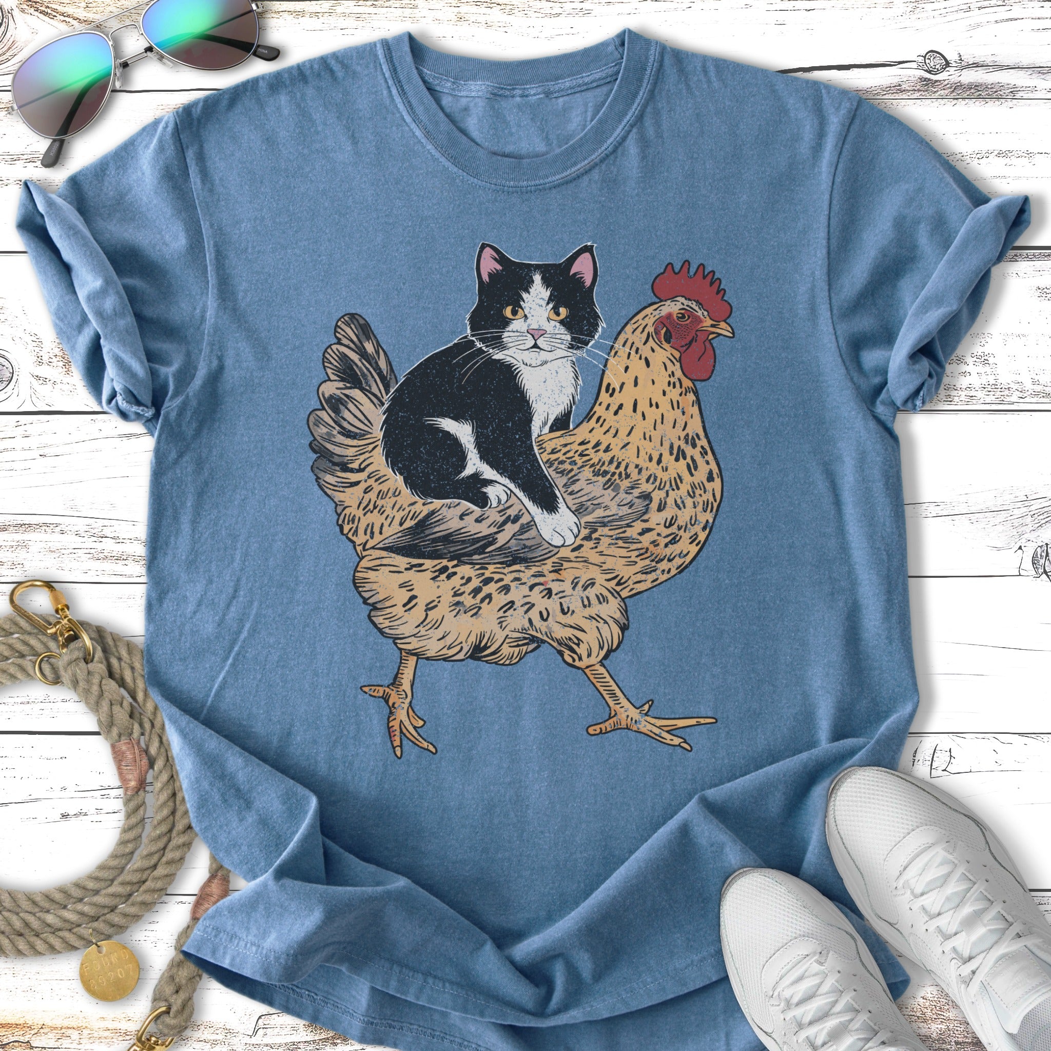 Cat sitting on a chicken graphic on a blue t-shirt.