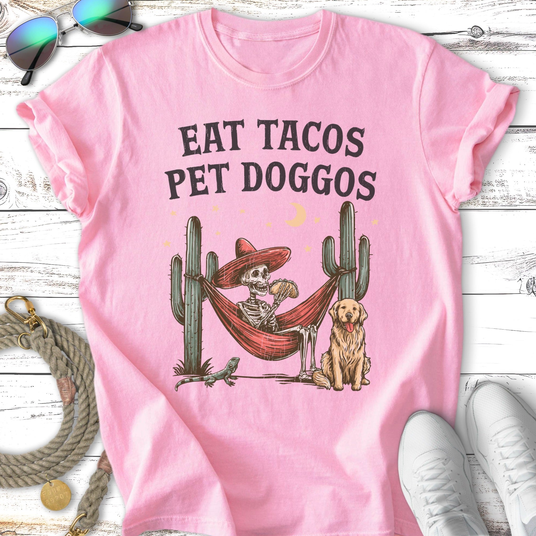 Pink t-shirt with "Eat Tacos Pet Doggos" text, skeleton in hammock with taco, dog, and cactus.