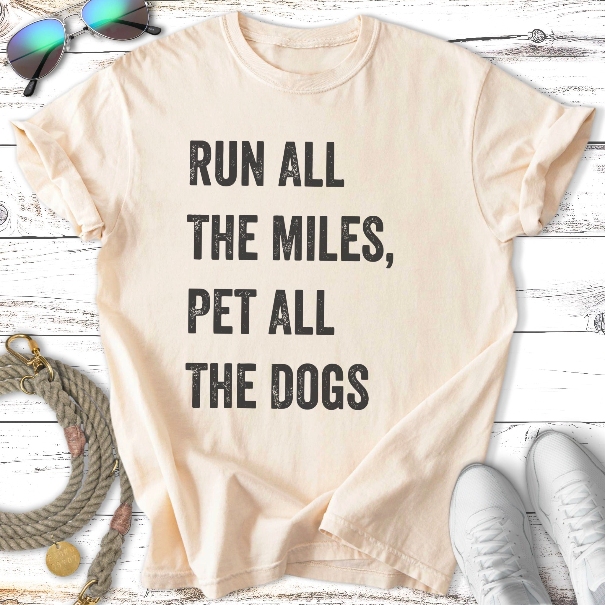 Cream t-shirt with "RUN ALL THE MILES, PET ALL THE DOGS" text, sunglasses, leash, and sneakers.