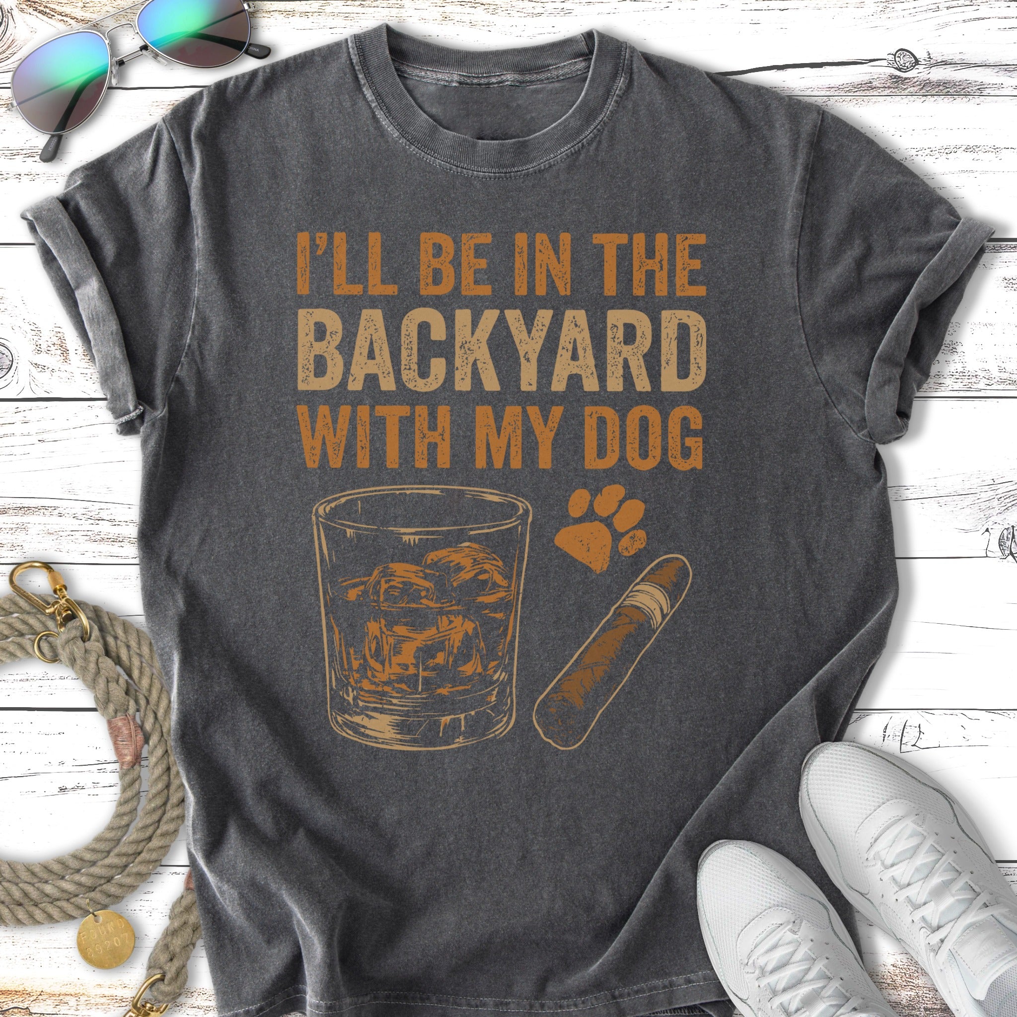 T-shirt with text "I'll be in the backyard with my dog," whiskey glass, and cigar.