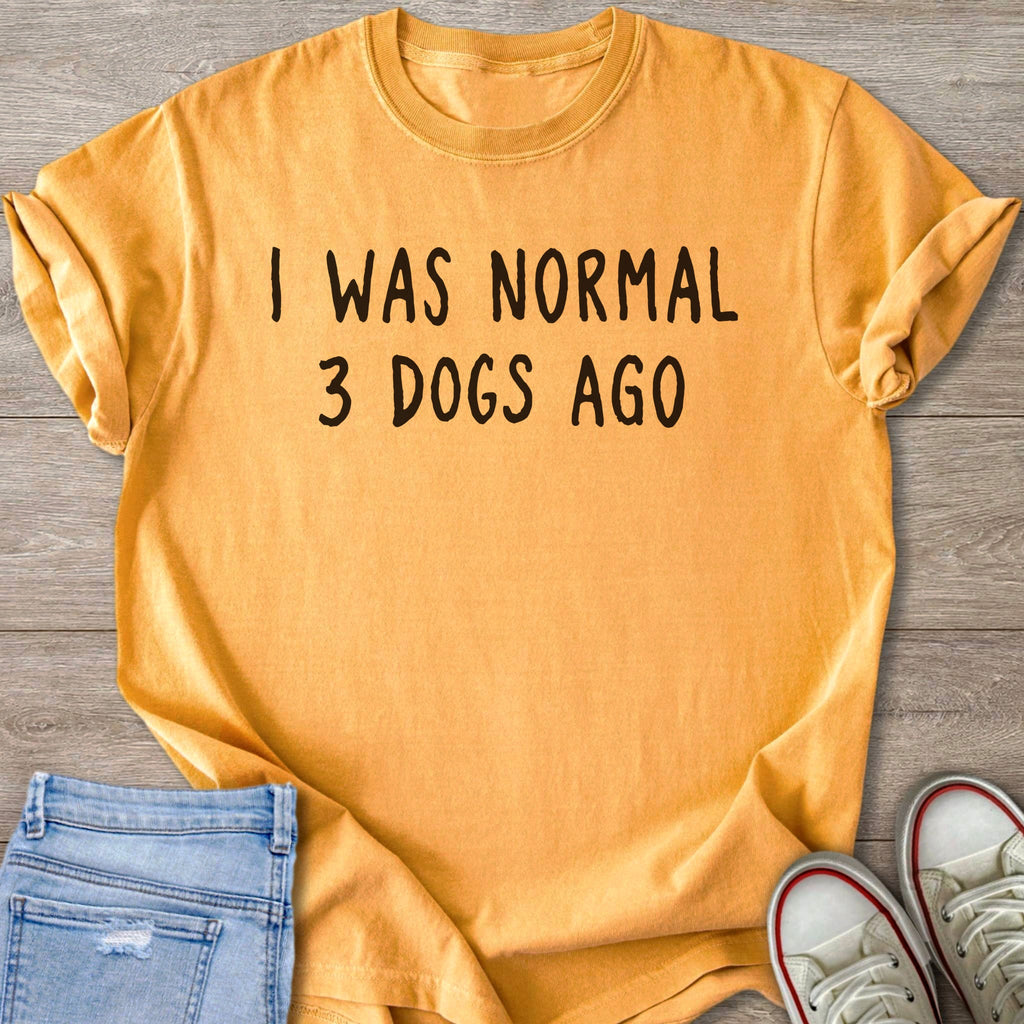Orange t-shirt with black text "I WAS NORMAL 3 DOGS AGO" on wooden background.
