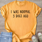 Orange t-shirt with black text "I WAS NORMAL 3 DOGS AGO" on wooden background.
