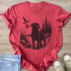 Black Labrador retriever standing in water with ducks flying and trees in background.