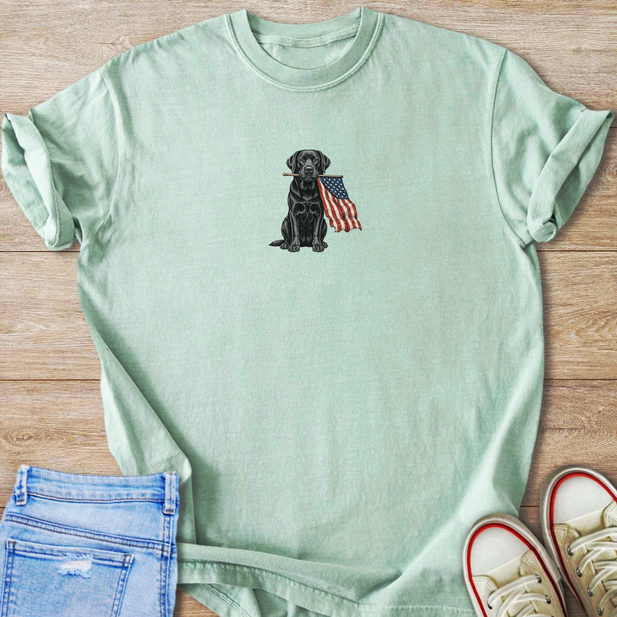 Black Labrador with American flag in mouth on a light green t-shirt.