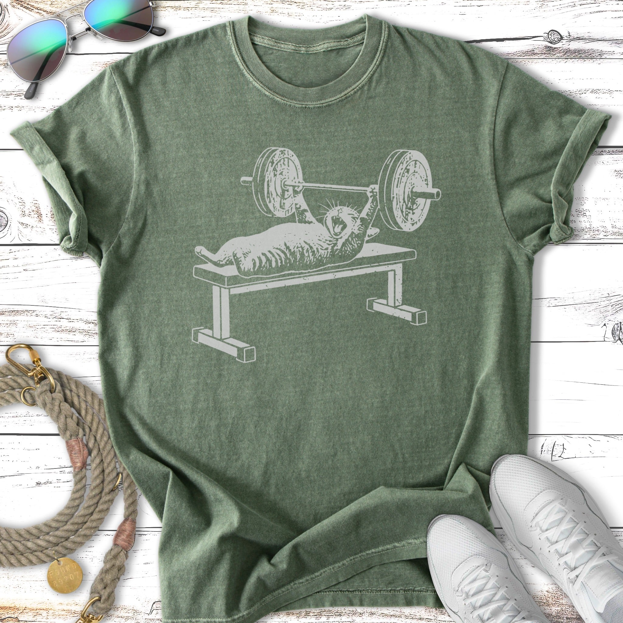 Cat lifting weights on a bench press, printed on a green t-shirt.