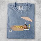 T-shirt featuring a dachshund wearing sunglasses, relaxing under a beach umbrella with a hot dog.