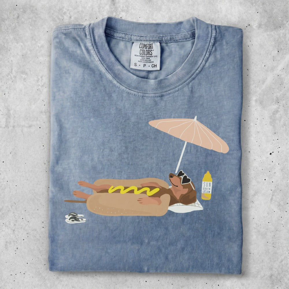 T-shirt featuring a dachshund wearing sunglasses, relaxing under a beach umbrella with a hot dog.