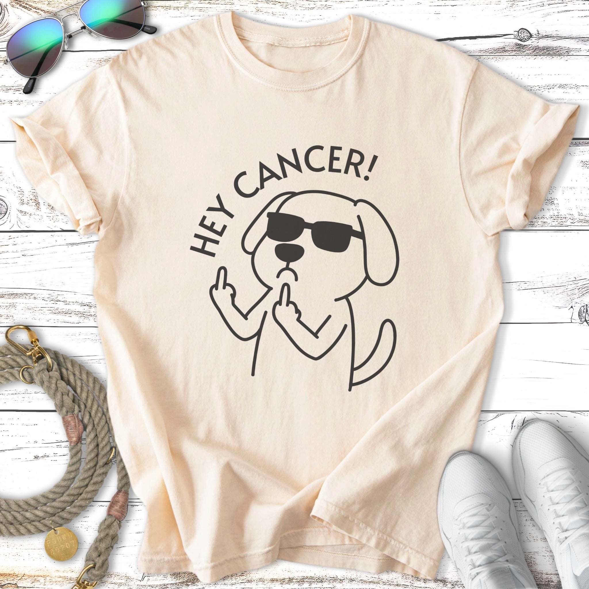 Cream t-shirt with black graphic of a dog wearing sunglasses flipping the bird and saying "Hey Cancer!".