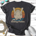 T-shirt graphic with fat cat being held by hands, sunset background, "Rolling Fatties" text.