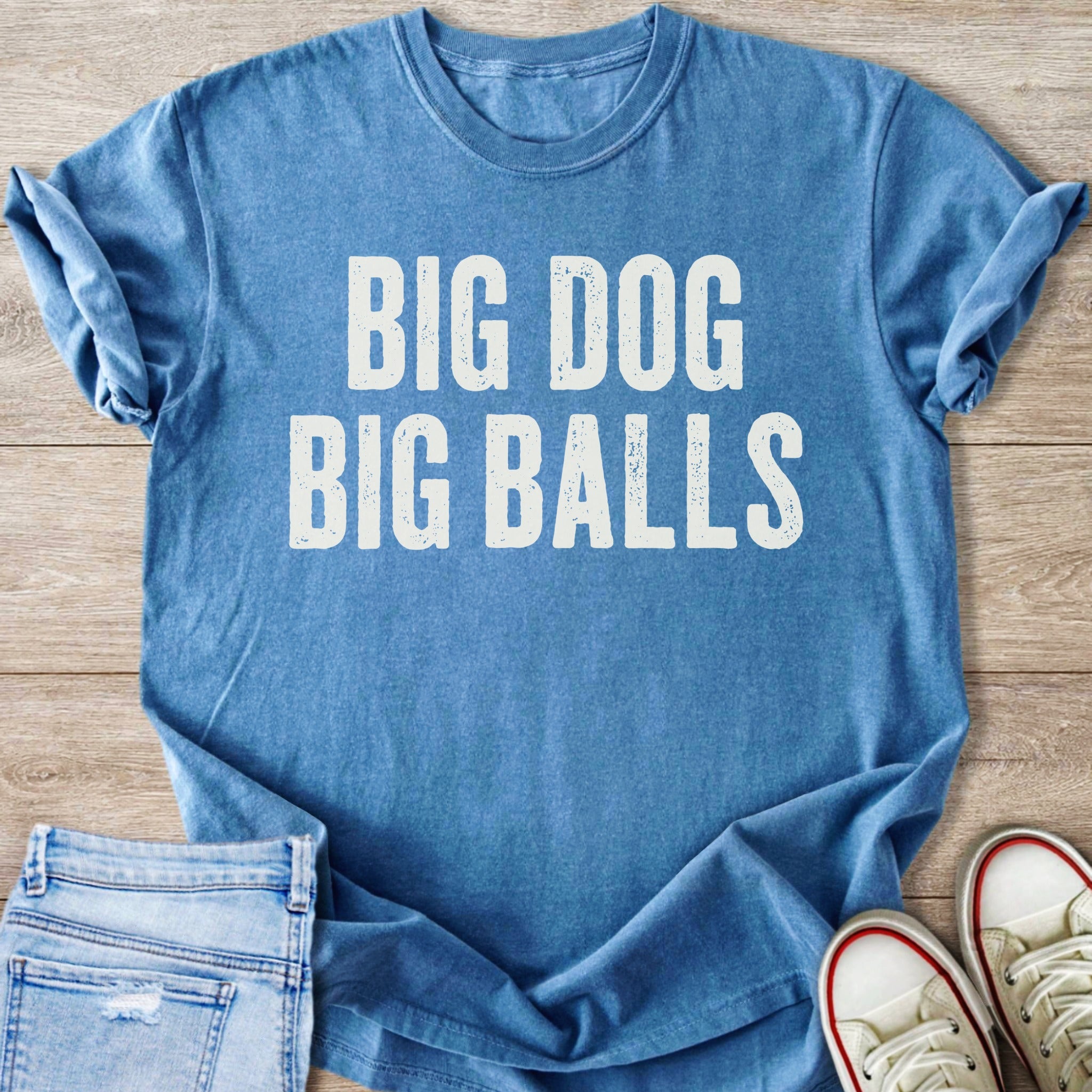 Blue t-shirt with white text "BIG DOG BIG BALLS" on a wooden surface.