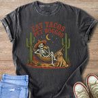 Skeleton in hammock eating taco with dog and cacti, text "EAT TACOS PET DOGGOS".