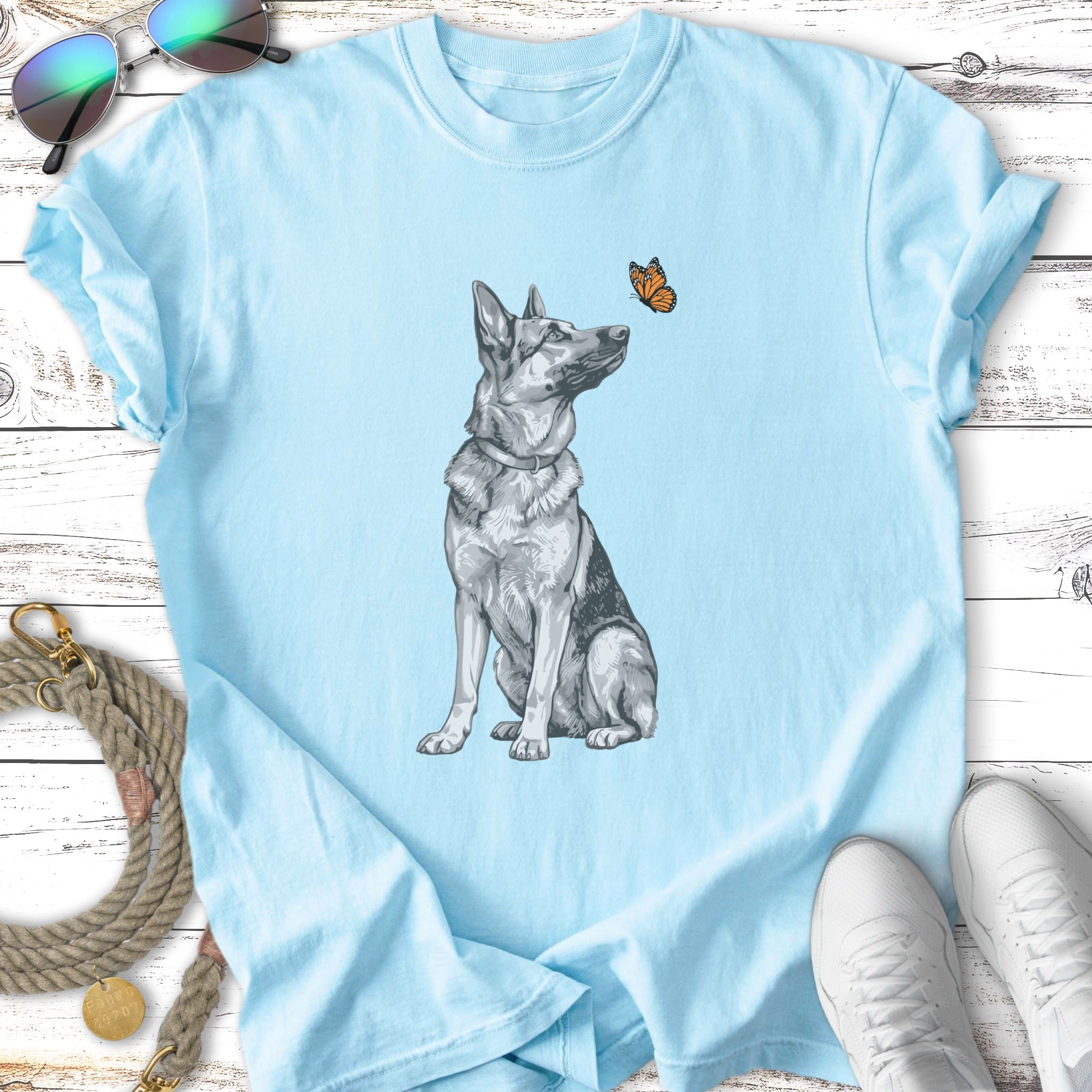 German Shepherd sitting, looking at a butterfly on a light blue t-shirt.