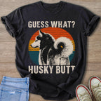 Black t-shirt on wooden background with husky graphic and text "GUESS WHAT? HUSKY BUTT".
