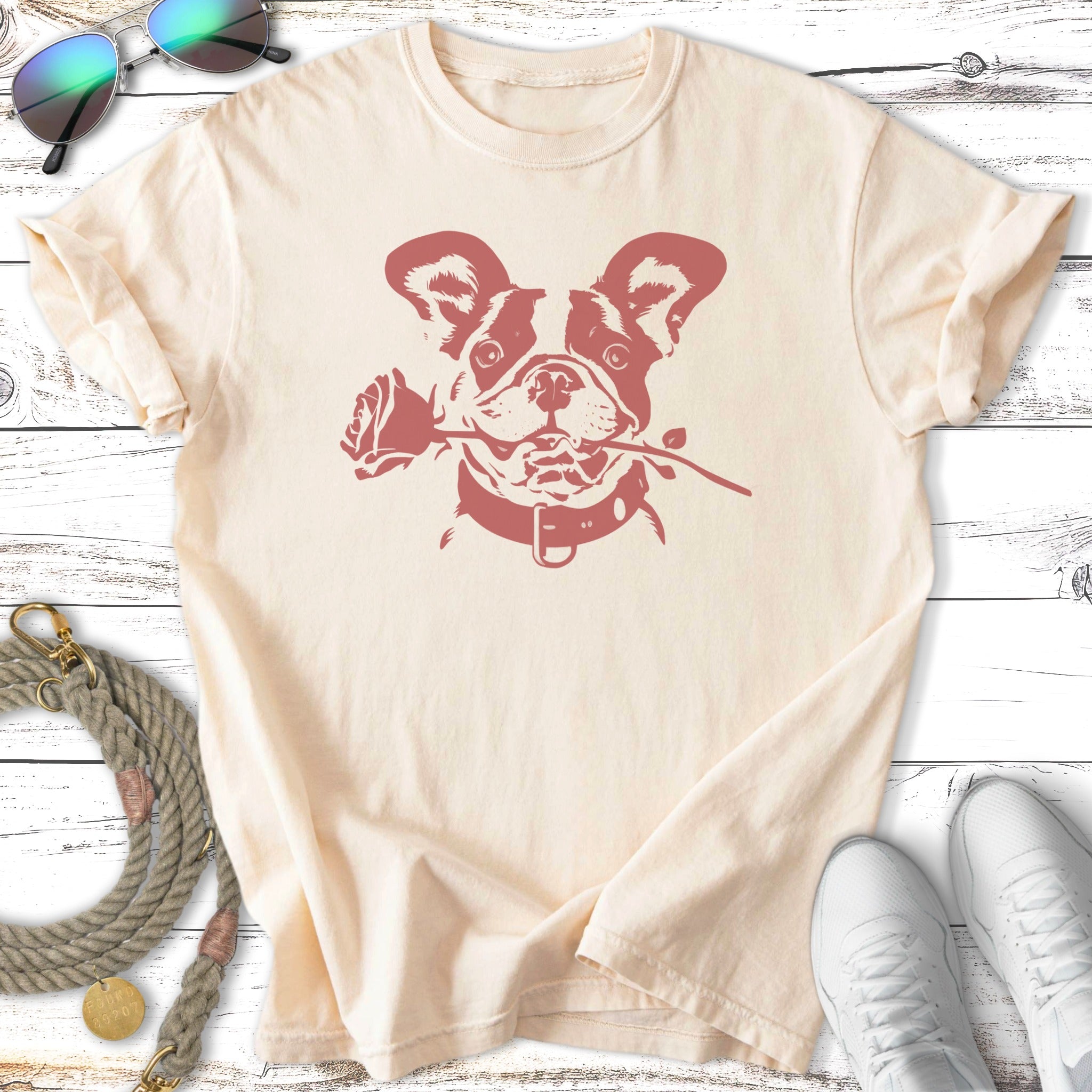 French bulldog wearing a collar holds a rose in its mouth on a cream t-shirt.