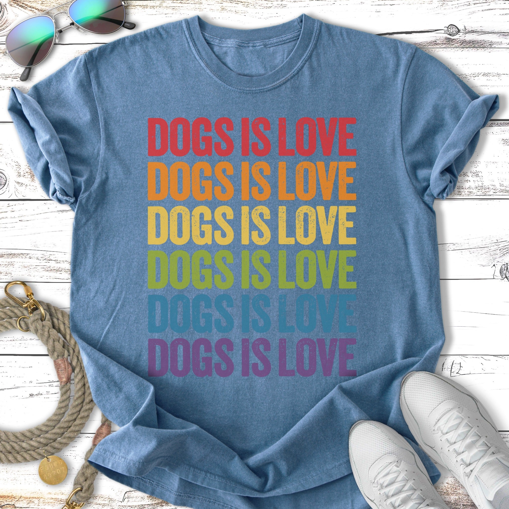 Blue t-shirt with "Dogs is Love" in rainbow colors, laying flat with sunglasses and shoes.