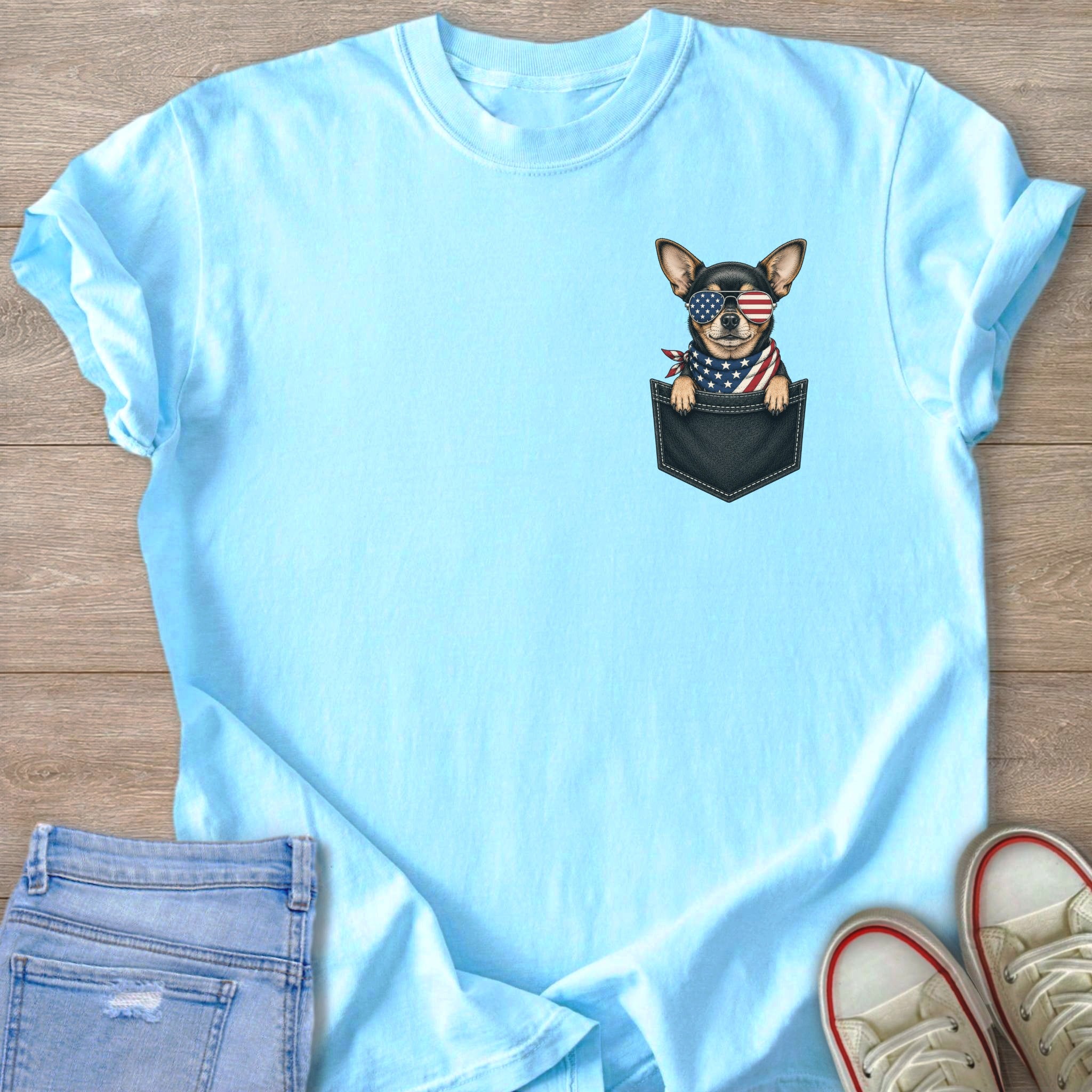 Chihuahua in pocket wearing American flag bandana and sunglasses on light blue t-shirt.