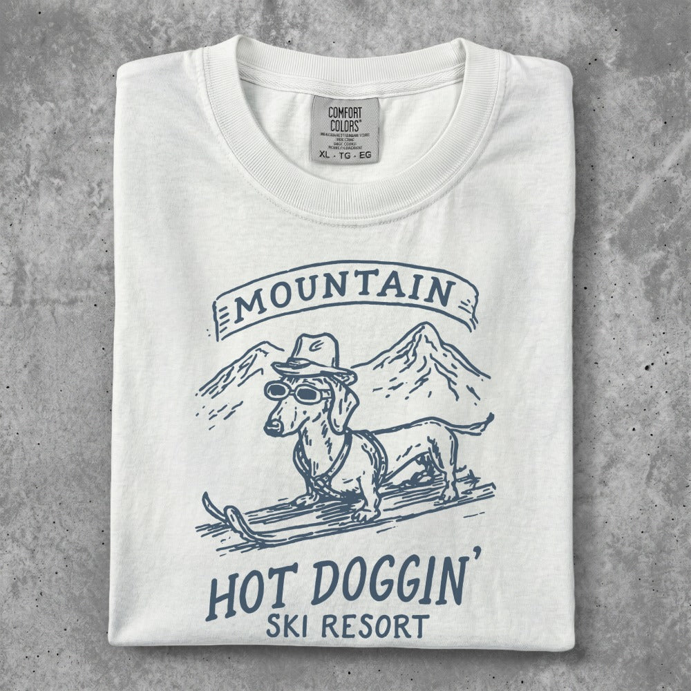 White t-shirt with graphic of a dachshund in sunglasses skiing, "Mountain Hot Doggin' Ski Resort" text.