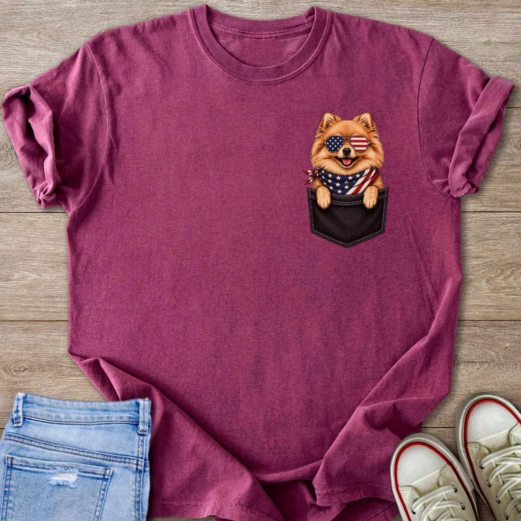 Pomeranian dog in patriotic sunglasses peeking from a pocket on a maroon t-shirt.