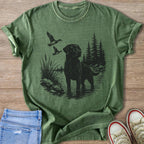 Black Labrador in water with ducks flying past pine trees.