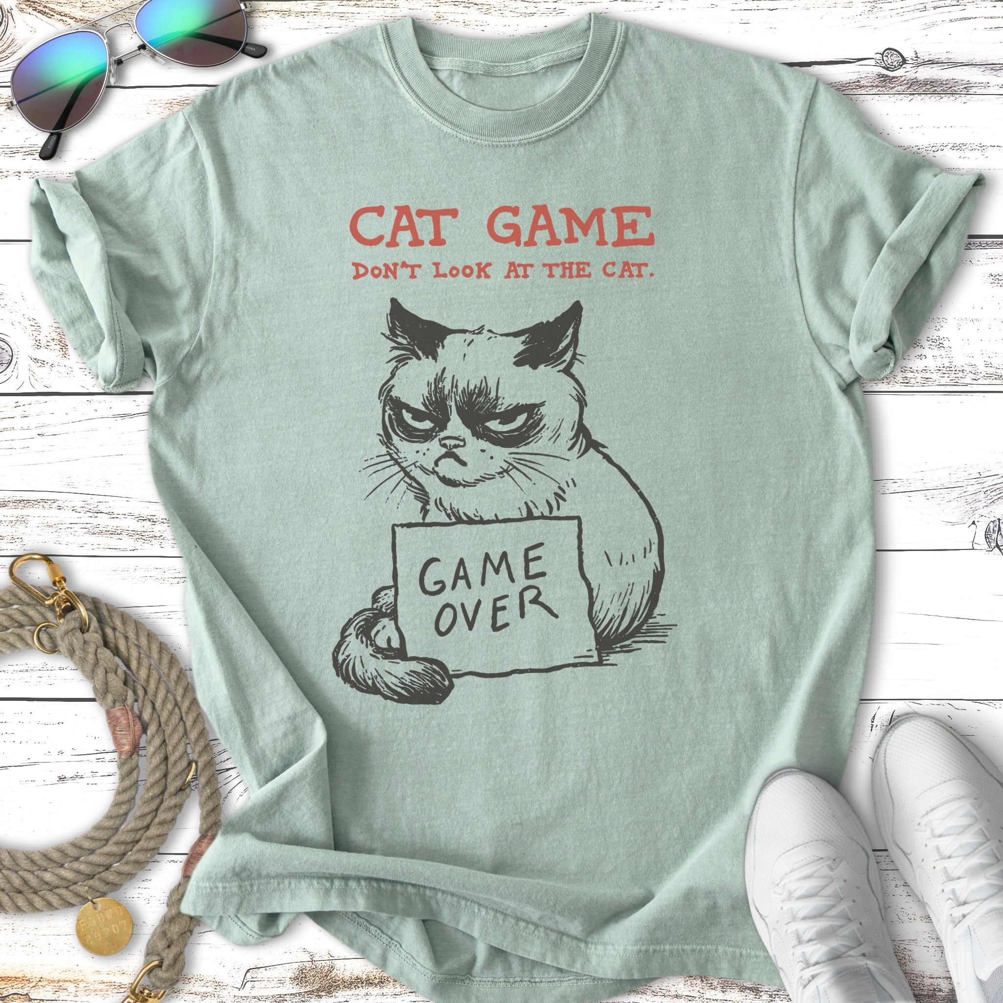 Green t-shirt with grumpy cat drawing, text "CAT GAME DON'T LOOK AT THE CAT. GAME OVER".