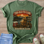 Green t-shirt with a graphic of a dog in a rocking chair by a lake, text "Too Old To Care Boomer Club".