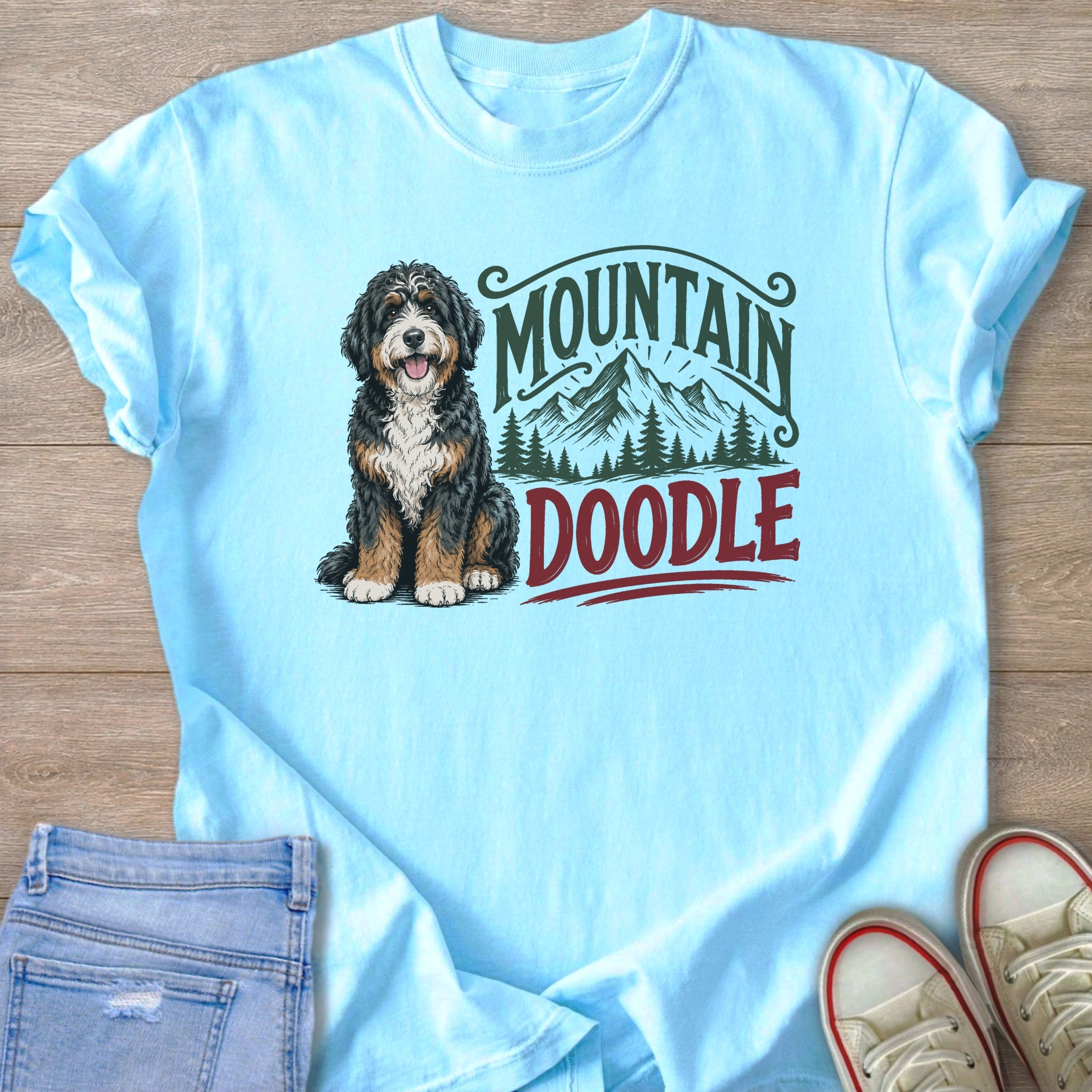 Bernedoodle dog with "Mountain Doodle" text and mountain graphics on light blue t-shirt.