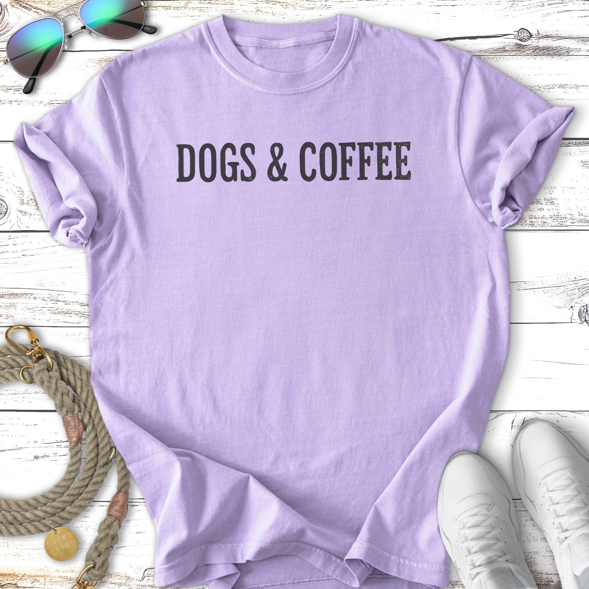 Lavender t-shirt with "DOGS & COFFEE" text, laid flat with sunglasses and sneakers.
