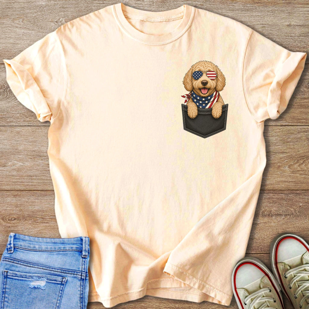 Peach t-shirt with a goldendoodle wearing American flag sunglasses and bandana peeking from pocket.