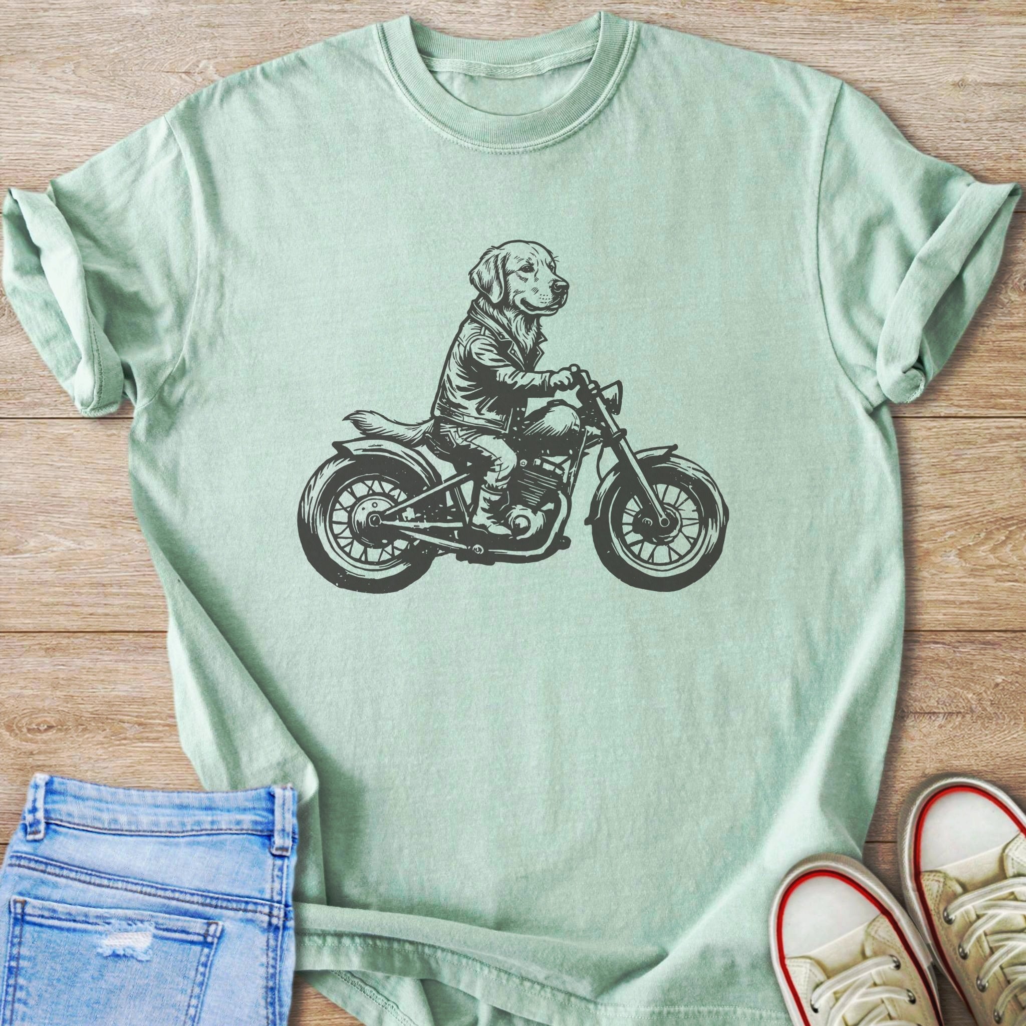 Line art of a dog wearing a jacket riding a motorcycle on a mint green t-shirt.