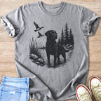 Black labrador stands in water with ducks flying over a wooded shoreline.