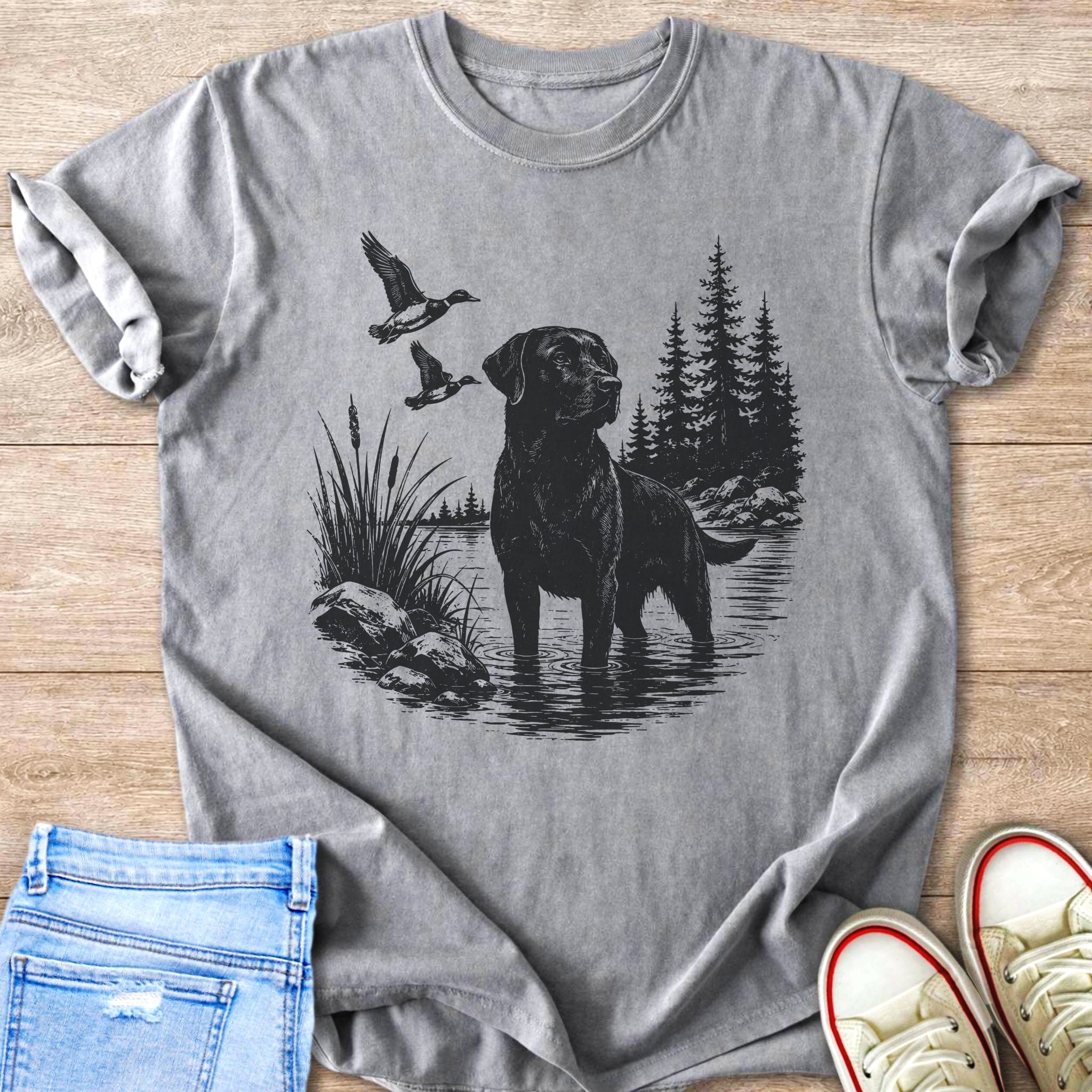 Black labrador stands in water with ducks flying over a wooded shoreline.