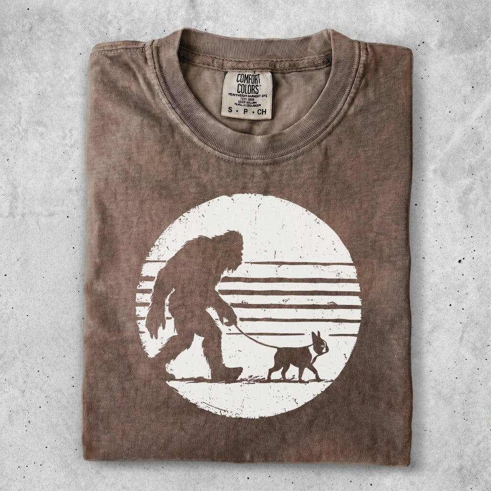 T-shirt with Bigfoot walking a dog graphic on a striped background.