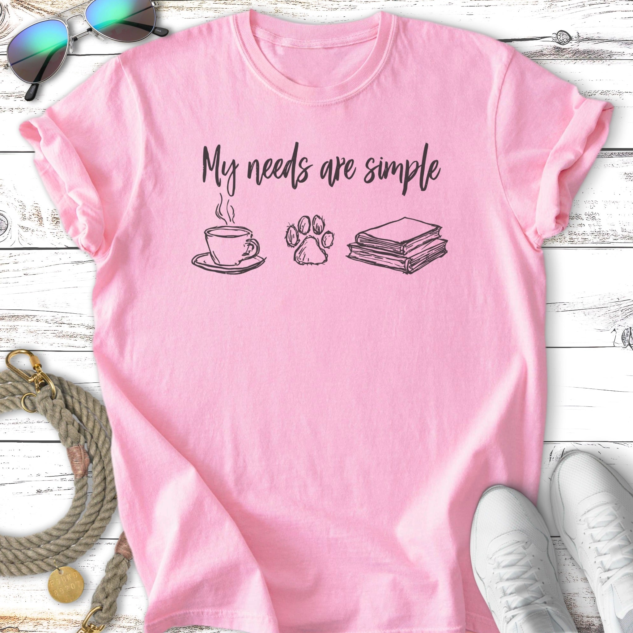 Pink t-shirt with "My needs are simple" and icons of coffee, paw print, and books.