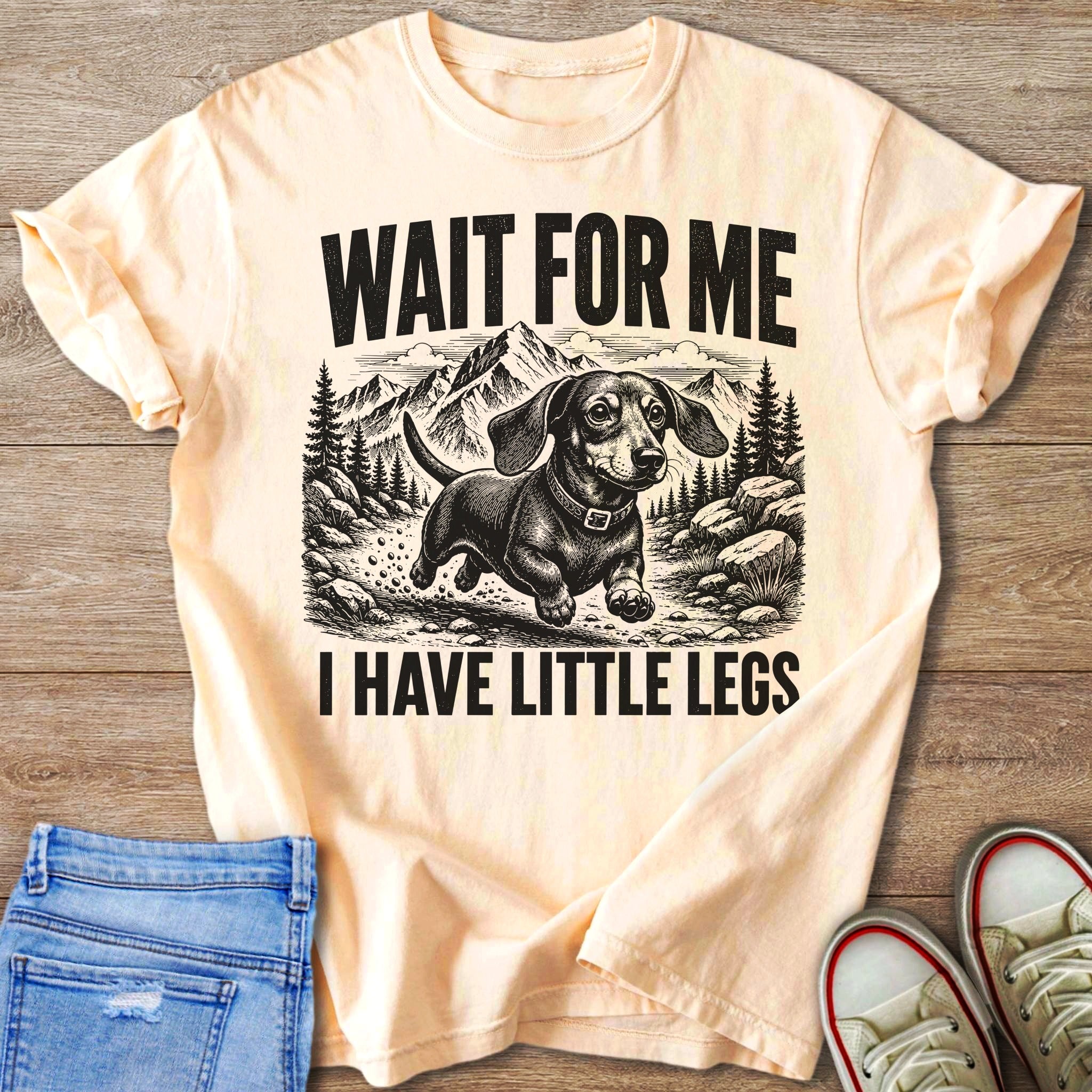 Peach t-shirt with black graphic of a dachshund running through mountains, text "WAIT FOR ME I HAVE LITTLE LEGS".