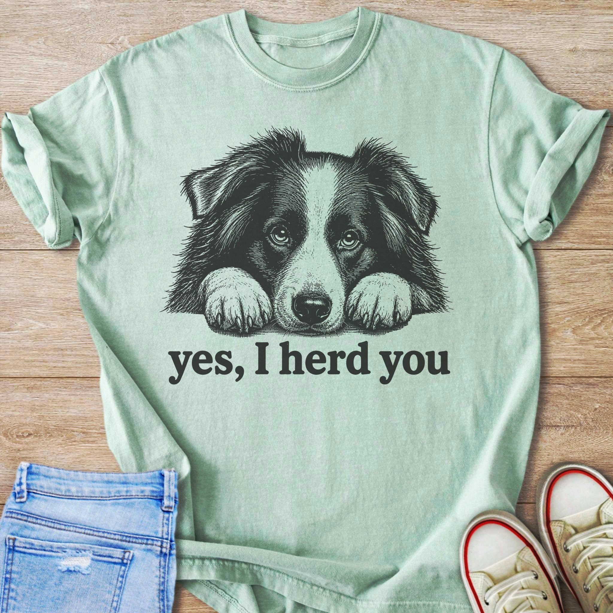 A black and white sketched border collie rests paws on text saying "yes, I herd you" on a light green t-shirt.