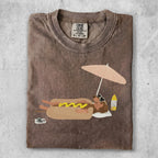 Brown t-shirt with cartoon dachshund wearing sunglasses, lying on beach towel under umbrella.