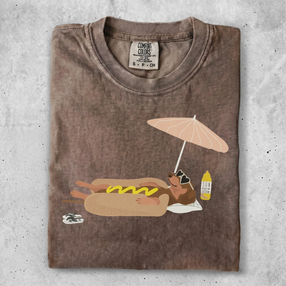Brown t-shirt with cartoon dachshund wearing sunglasses, lying on beach towel under umbrella.