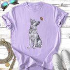 German Shepherd dog sits, looking up at a butterfly on a purple t-shirt.