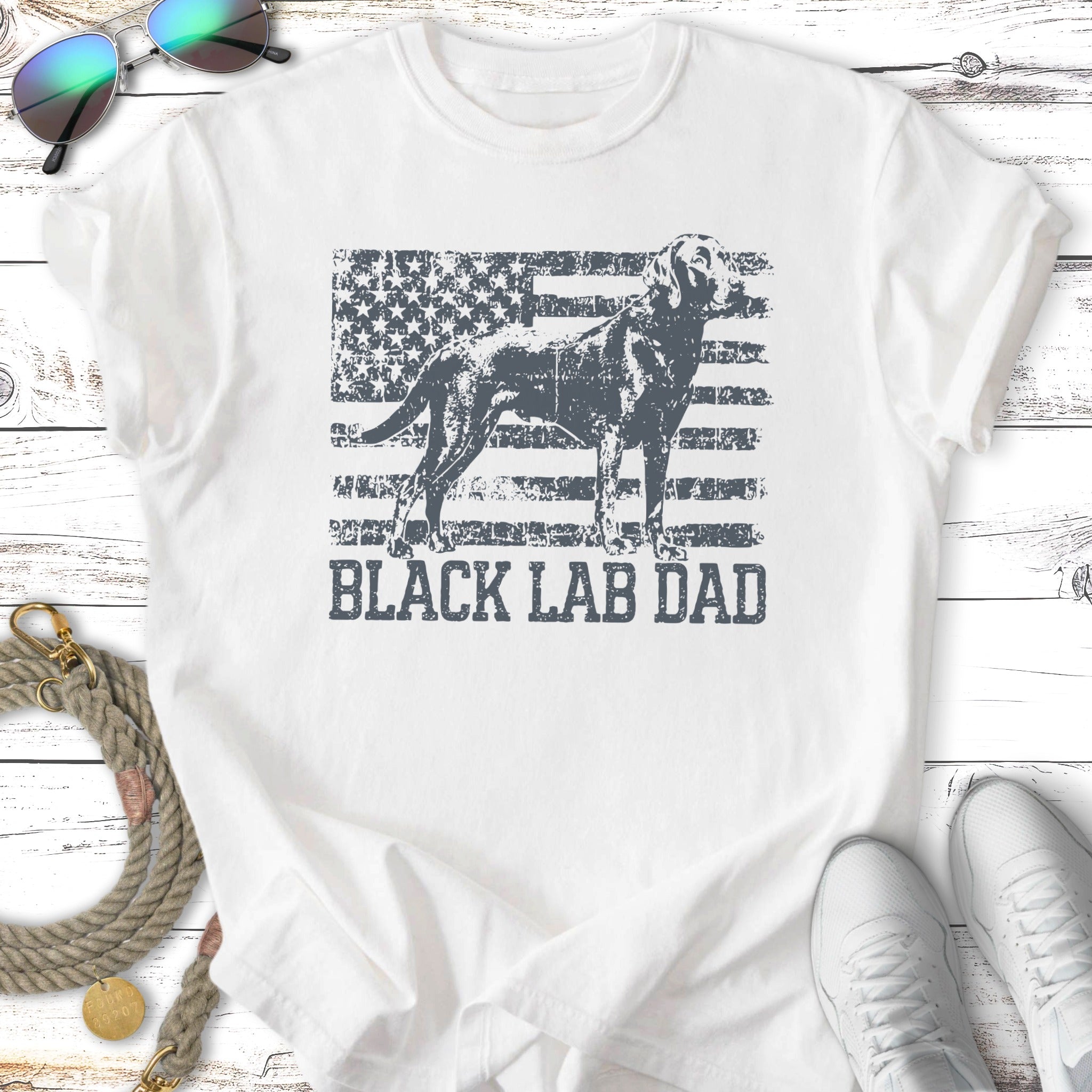 Black Labrador dog standing against distressed American flag graphic on white t-shirt.