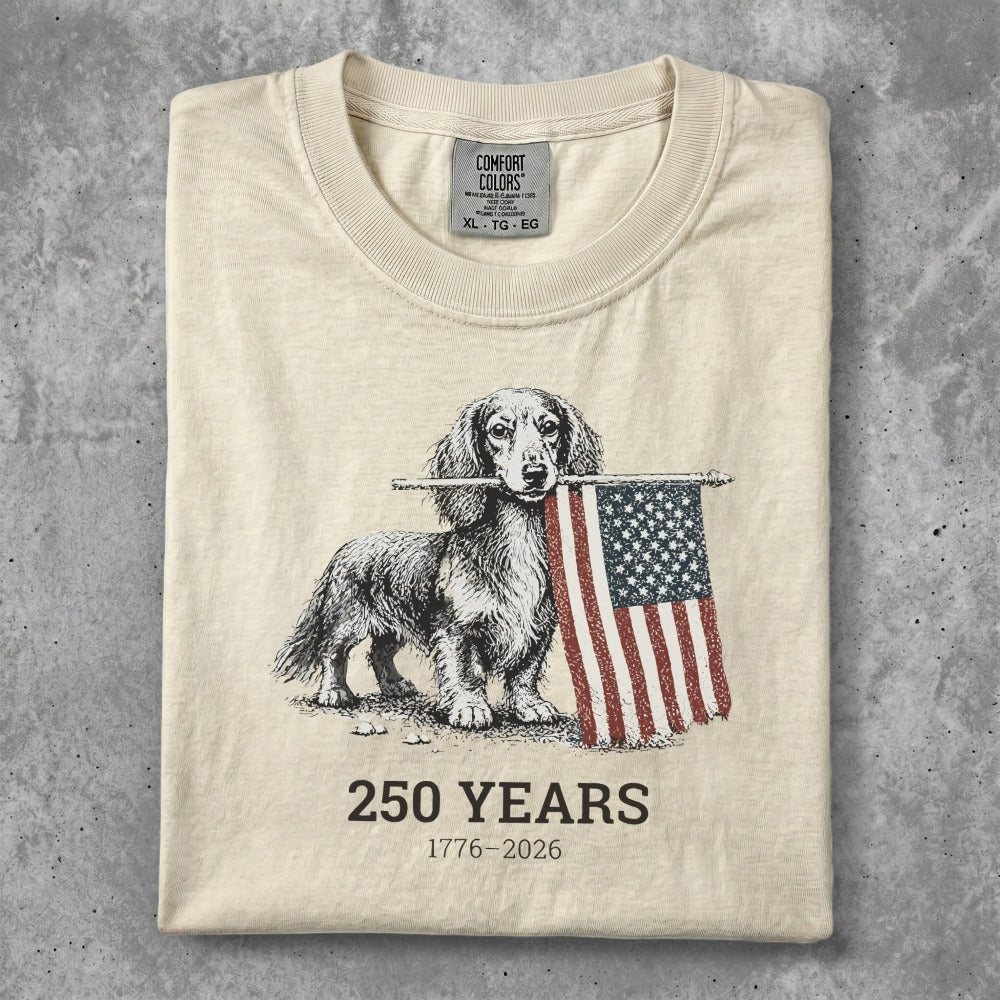 Dachshund holding American flag, "250 Years 1776-2026" text, cream t-shirt.