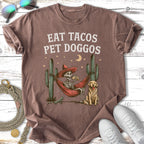 T-shirt with skeleton in hammock, dog, cactus, and text "EAT TACOS PET DOGGOS".